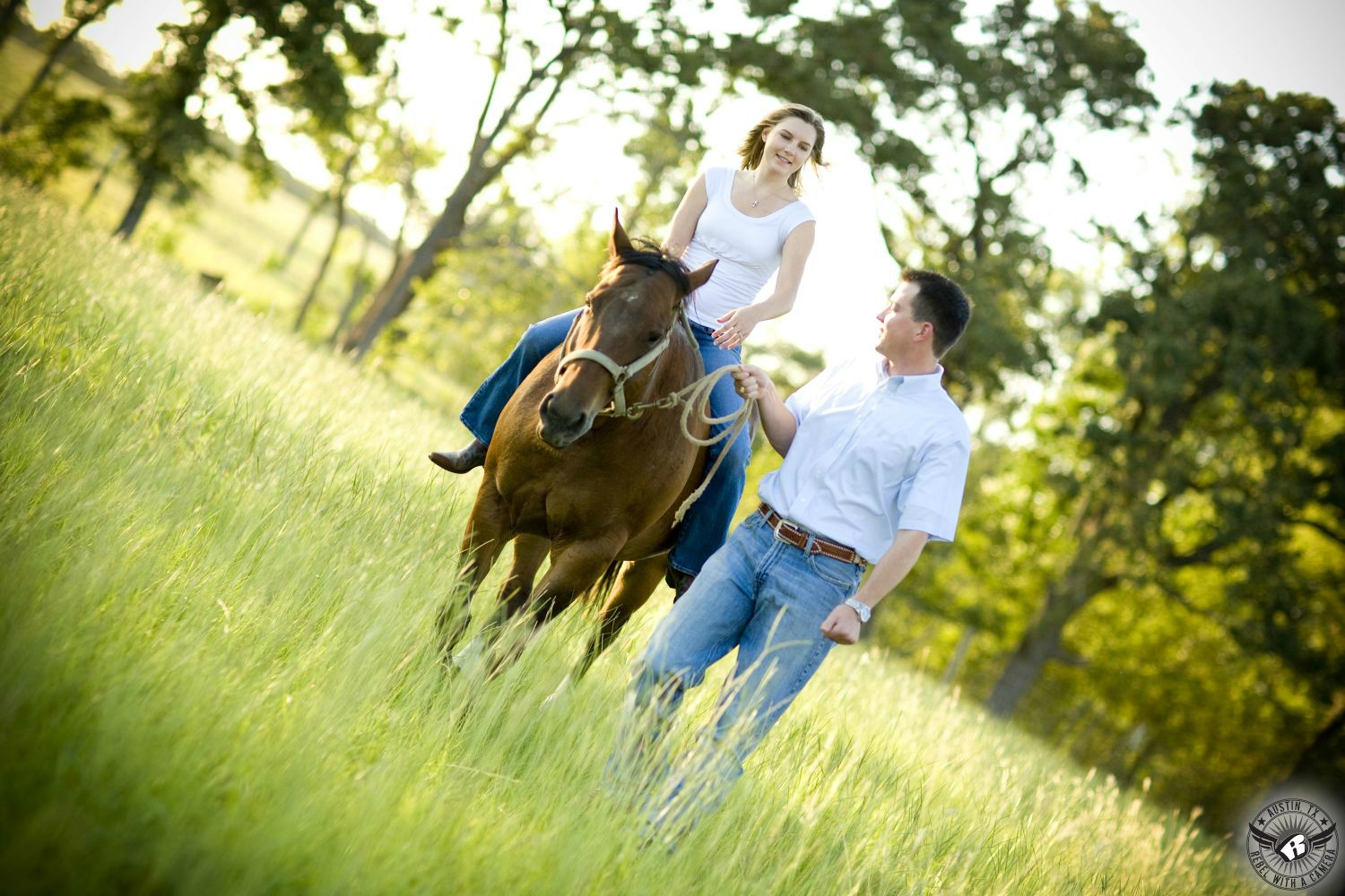 blond girl with white tank top and blue jeans ride brown horse being led by a guy with brown hair wearing a light blue button up shirt and blue jeans while walking through a filed of tall gold and green grass with trees out of focus in the back ground of this engagement photo